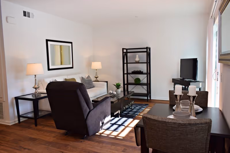 A bright living room with wooden flooring featuring a white sofa with cushions, a dark armchair, two side tables with lamps, a black shelving unit with decorative items, a flat-screen TV on a stand, and a dining table set with plates, bowls, and candles near a sliding glass door.
