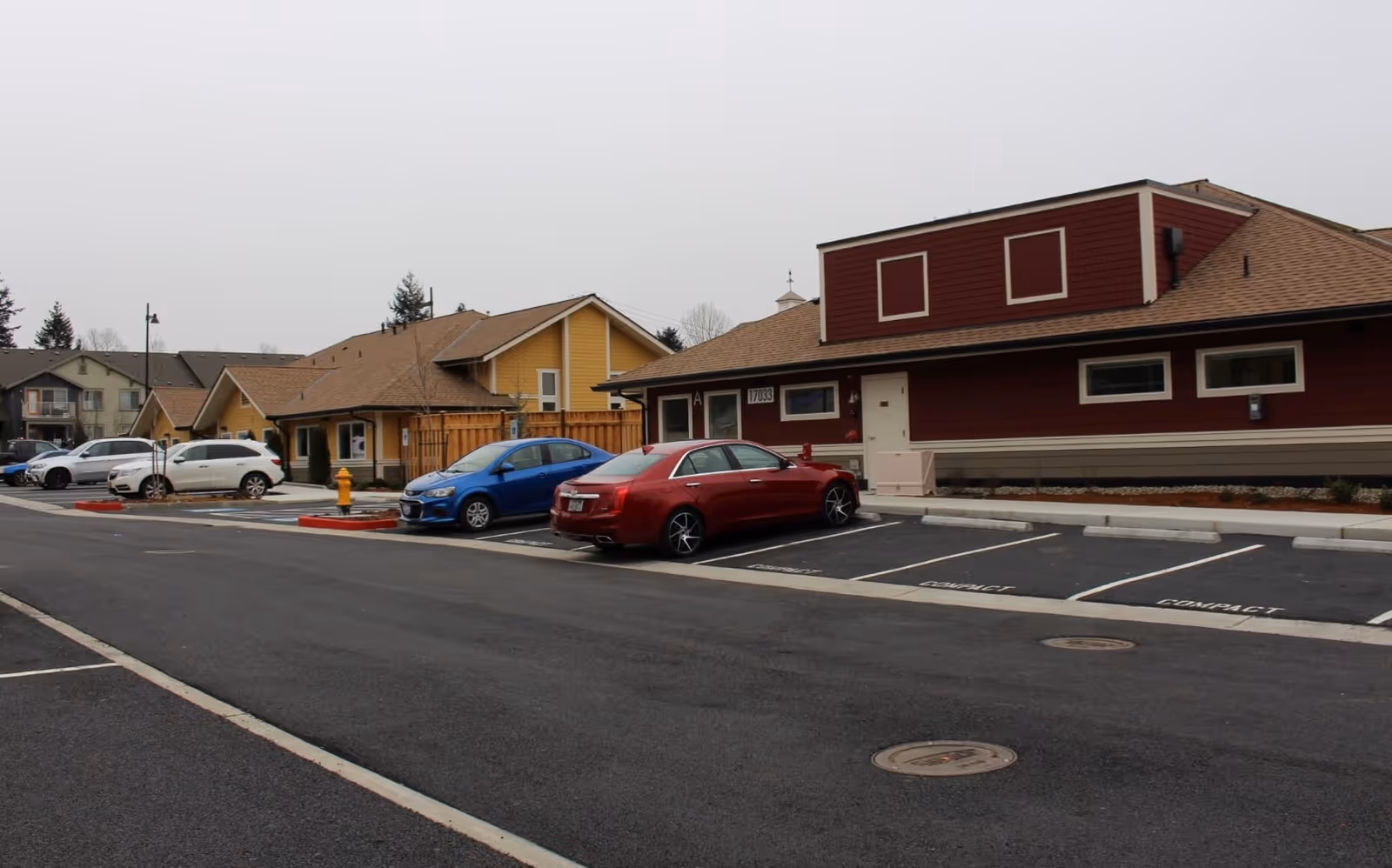 Parking lot in front of single-story cottage-style buildings with several parked cars under an overcast sky.