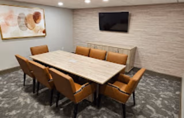 A modern meeting or dining room with a rectangular wooden table surrounded by eight brown upholstered chairs. The room has a carpeted floor, a wall-mounted flat screen TV, a wooden cabinet beneath the TV, and abstract artwork on one wall.