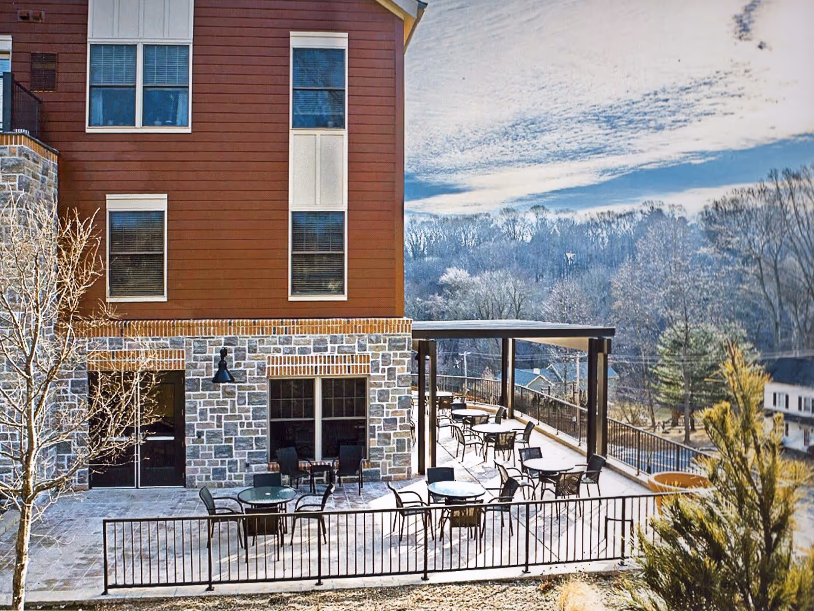 Outdoor patio area at Plush Mills Senior Living with multiple round tables and chairs arranged on a stone-paved terrace. The patio is adjacent to a building with stone and wood siding, and there is a pergola providing partial shade over some tables. Trees and a partly cloudy sky are visible in the background.