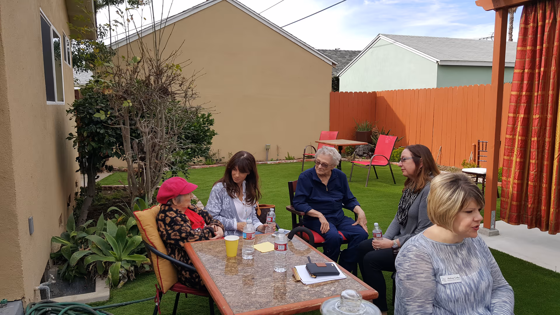 Five women sitting and talking around a rectangular outdoor table on a green lawn in a fenced backyard area with houses in the background. The table has water bottles, a yellow cup, and some papers on it. One woman is wearing a pink hat and a patterned jacket, while another woman has a name tag that reads 'Melissa Colby'.