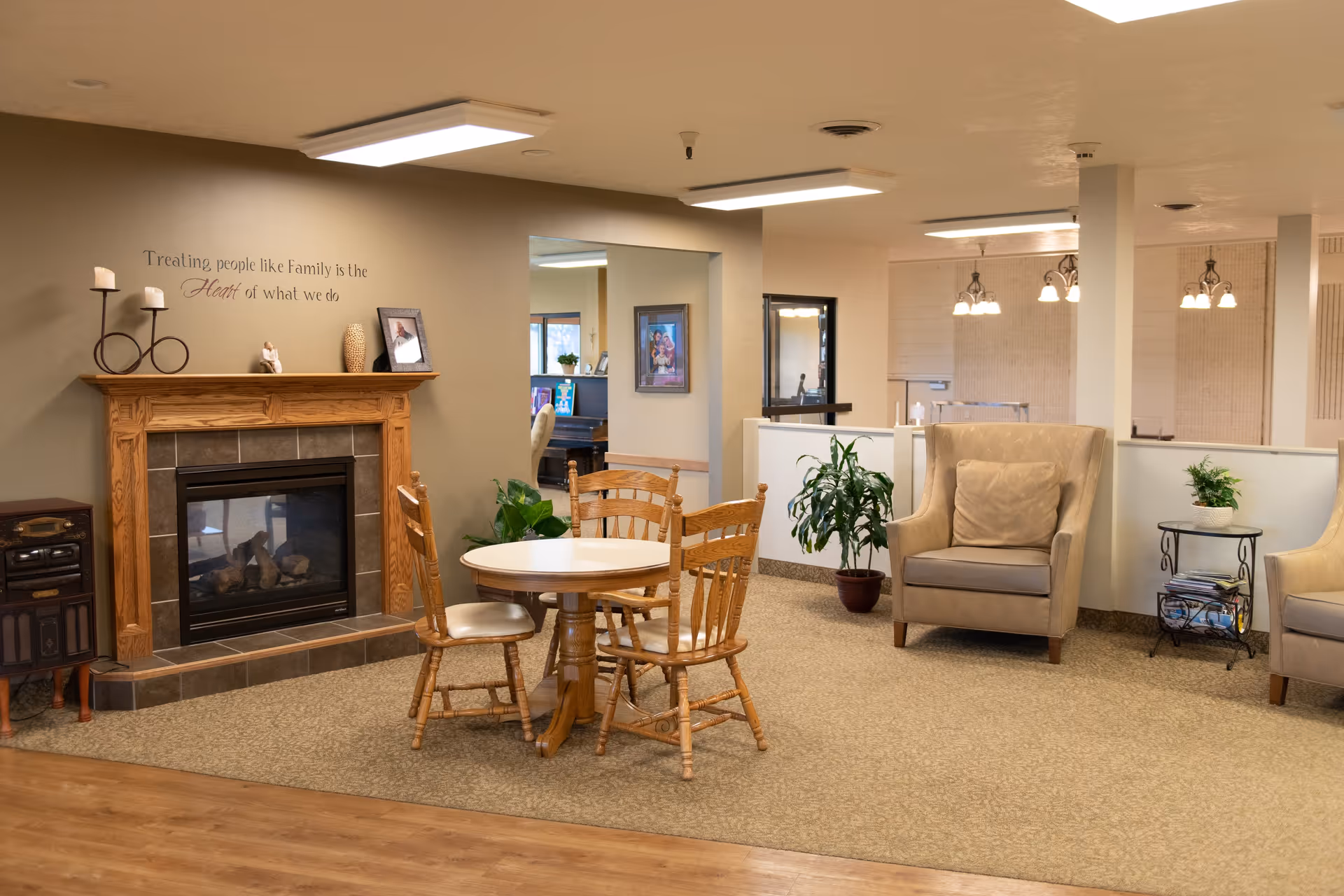 A cozy common area in a senior living facility featuring a wooden fireplace with decorative items on the mantel, a round table with four wooden chairs, two beige armchairs with cushions, potted plants, and soft lighting. The wall above the fireplace has a quote that reads, 'Treating people like Family is the Heart of what we do.'