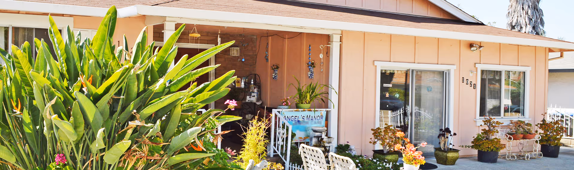 Exterior view of a single-story peach-colored house with a covered porch. The porch has various potted plants, hanging decorations, and a sign that reads 'Angel's Manor Care.' There are large green leafy plants and flowers in the front yard, along with white patio chairs and a small fountain. The house has sliding glass doors and windows with white trim.