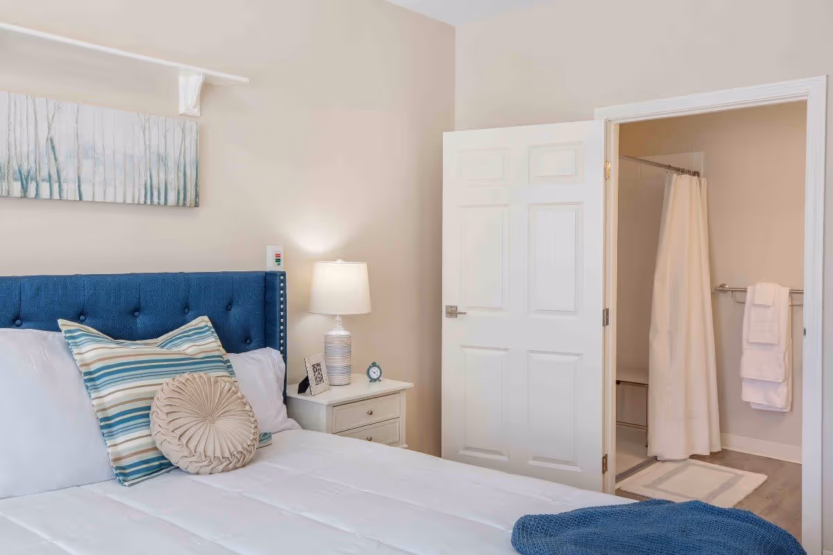 A bedroom with a blue upholstered headboard, white bedding, and two decorative pillows. A white nightstand with a lamp, a small clock, and a framed photo is beside the bed. An open door reveals a bathroom with a shower curtain, towel rack with white towels, and a bath mat on the floor.