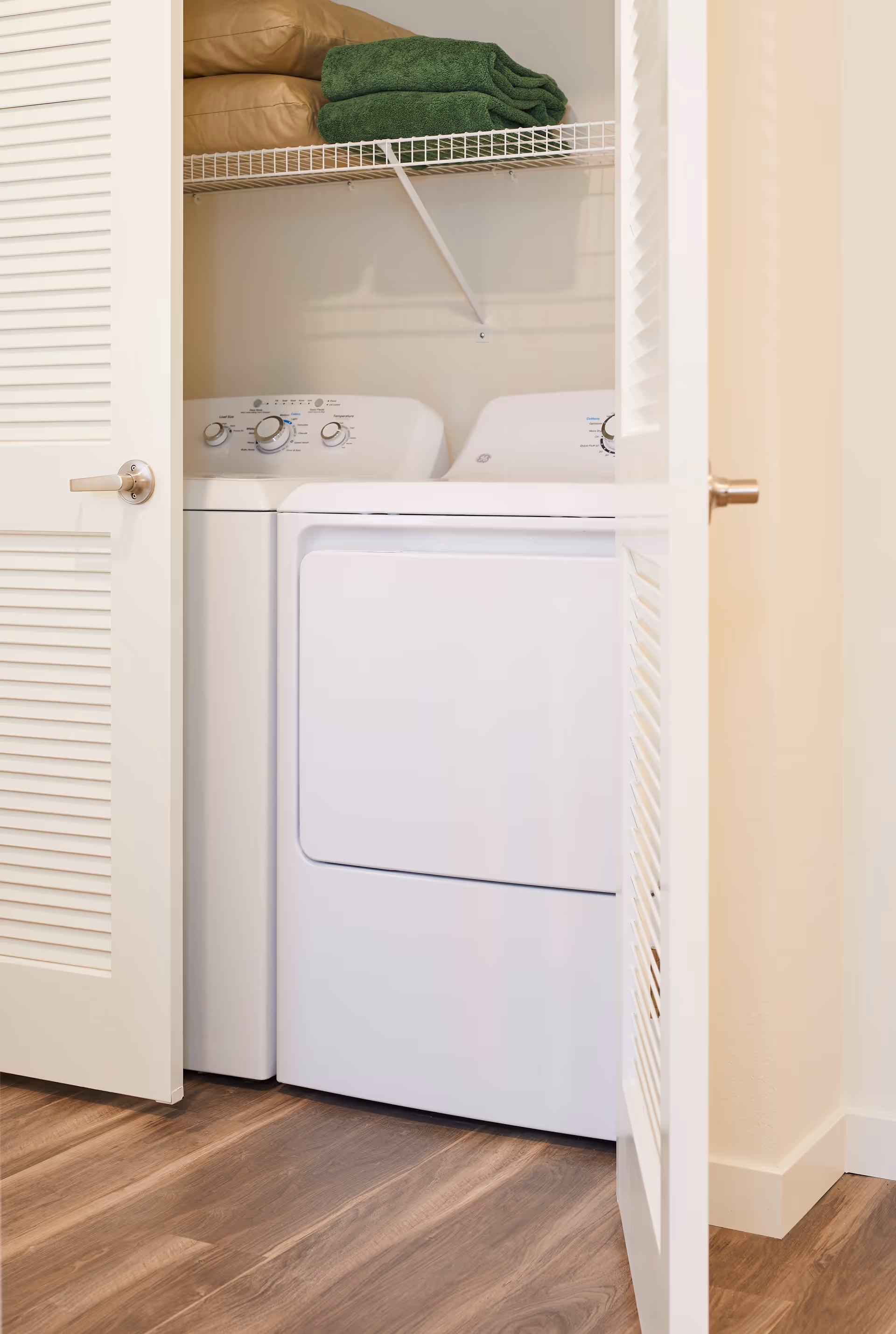 A laundry closet with white washing machine and dryer side by side, partially open white louvered doors, and a wire shelf above holding folded green towels and a beige cushion.