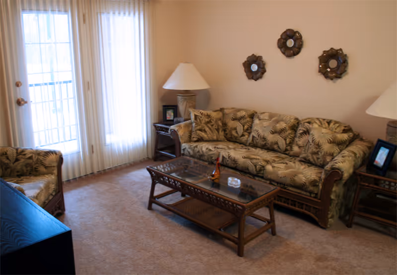 A cozy living room with a patterned sofa and matching armchair, a glass-top coffee table, two side tables with lamps, and three decorative round mirrors on the wall. Light filters in through sheer curtains covering a door and window.
