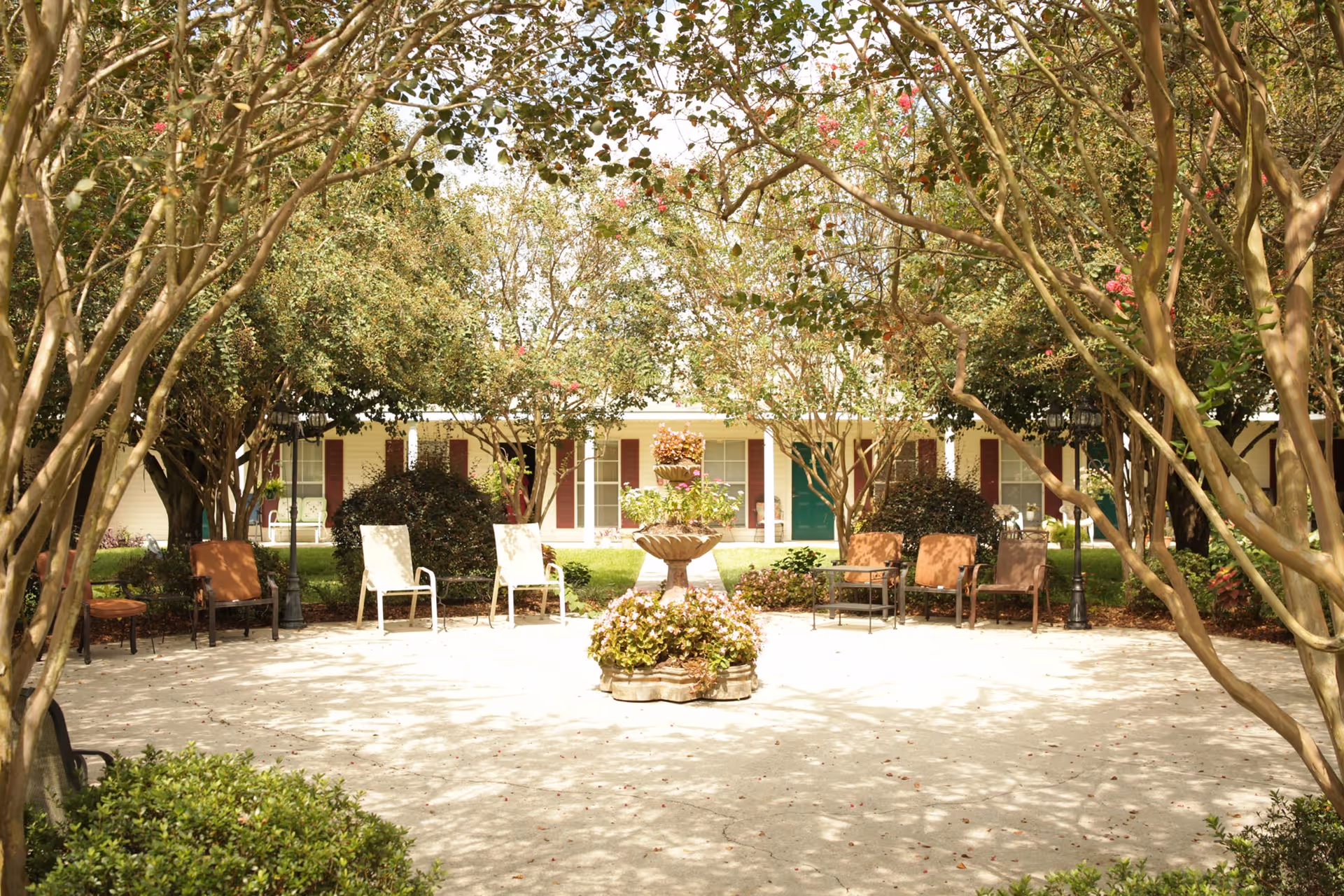 Shaded courtyard with chairs and a central planter/fountain framed by trees in front of a single-story residential building.