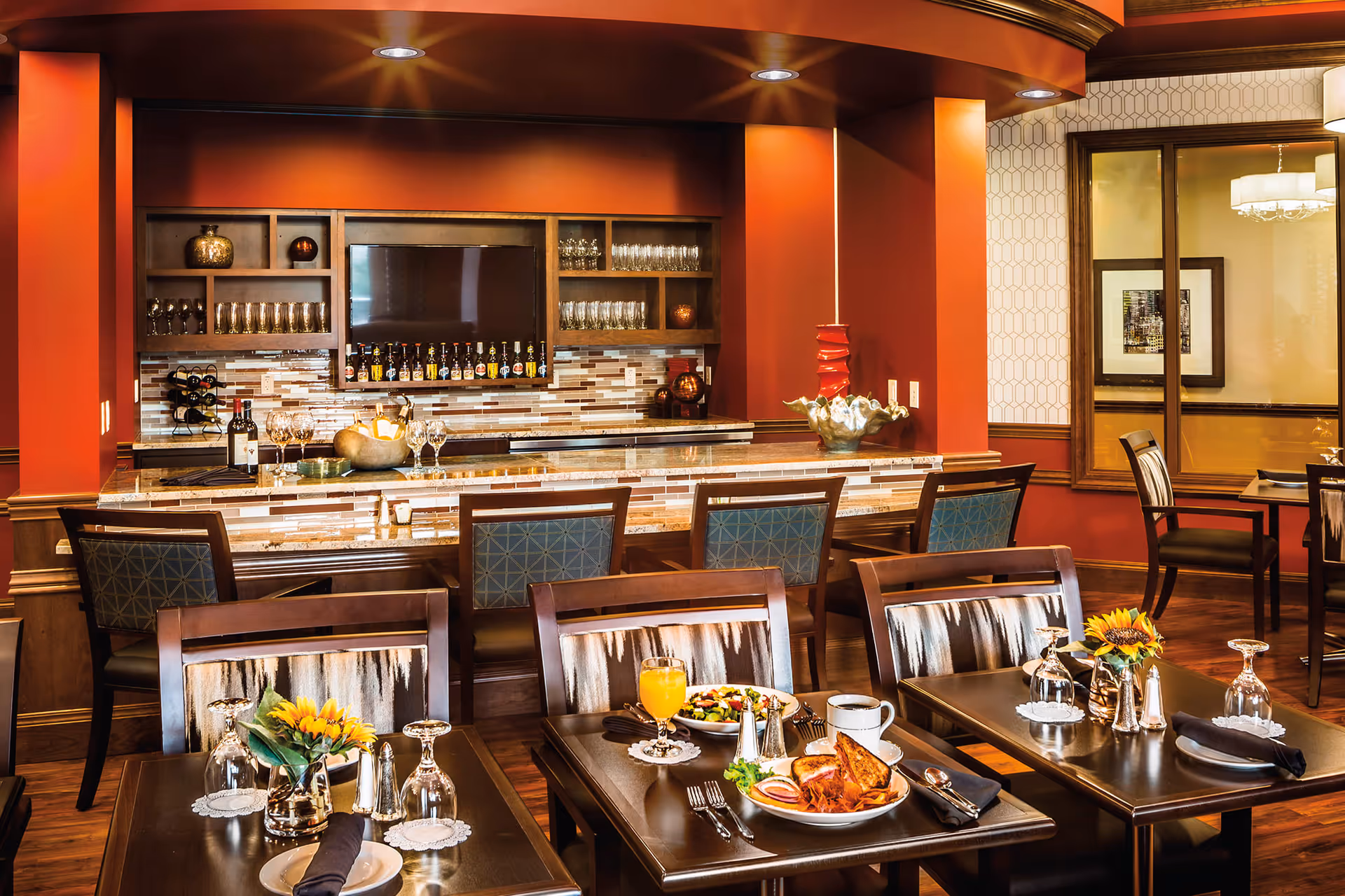 A warm and inviting dining area with wooden tables and chairs, each table set with glassware, napkins, and small flower arrangements. In the background, there is a bar with a marble countertop, shelves stocked with glasses and bottles, and a large flat-screen TV mounted on the wall. The walls are painted in a rich red color, and the room features wood paneling and flooring.