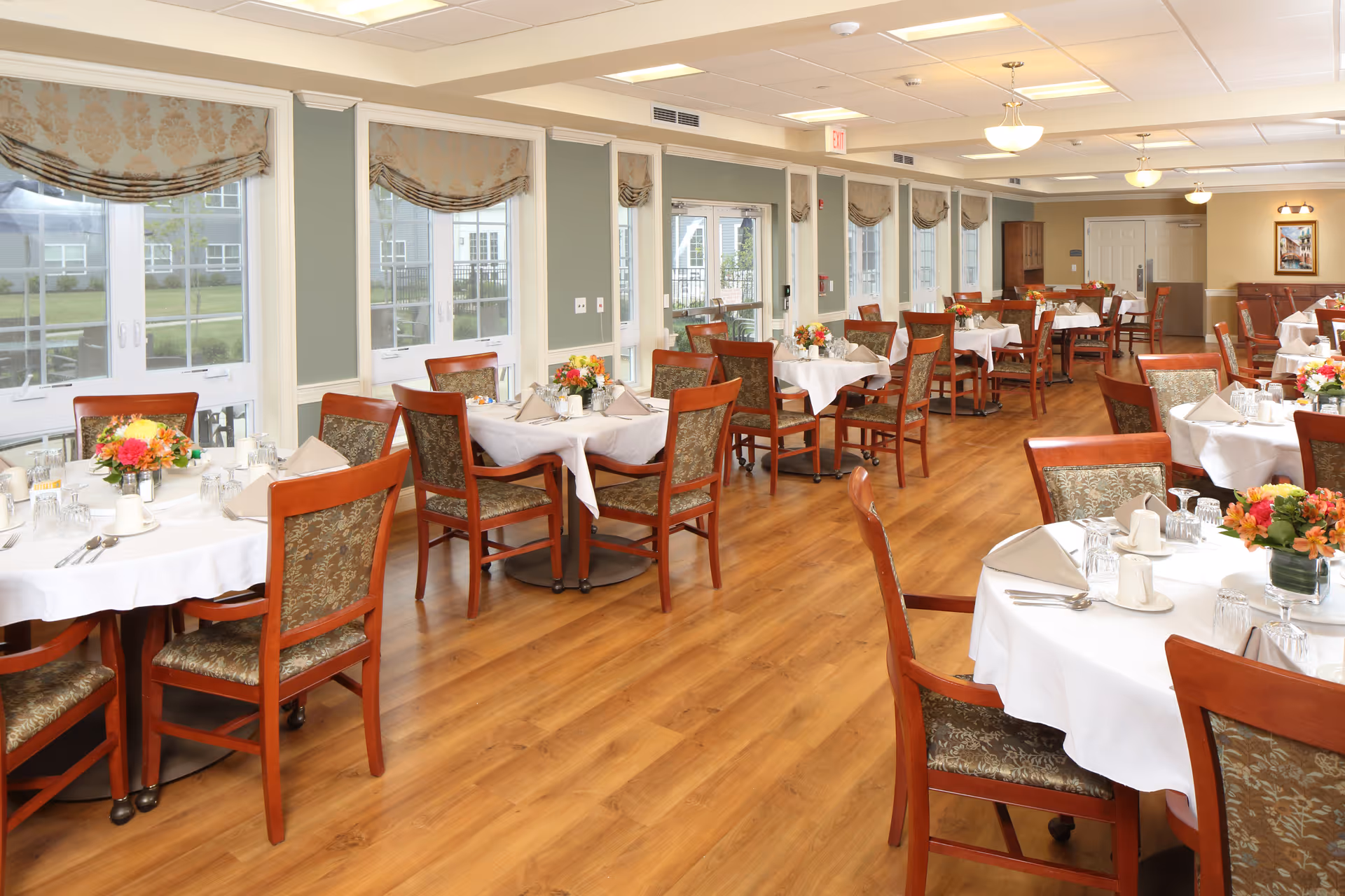 A bright and spacious dining room with multiple round tables covered in white tablecloths, each set with napkins, glasses, and floral centerpieces. The room has wooden chairs with patterned cushions, large windows with decorative valances, and wood flooring.