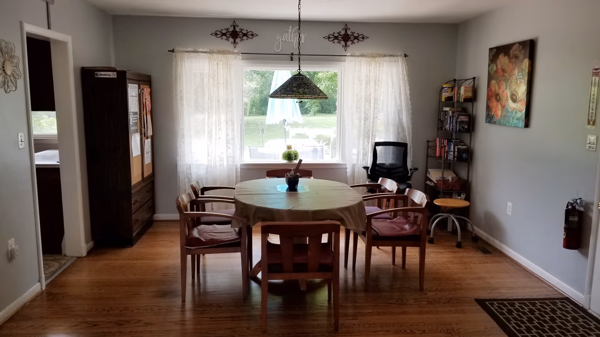 A dining room with a round table covered with a beige tablecloth surrounded by six wooden chairs with cushions. A hanging stained glass lamp is above the table. There is a large window with sheer white curtains letting in natural light and showing a green outdoor area with a patio umbrella. The room has wooden floors, a small bookshelf with books and games, a black office chair, a fire extinguisher mounted on the wall, and decorative wall art.