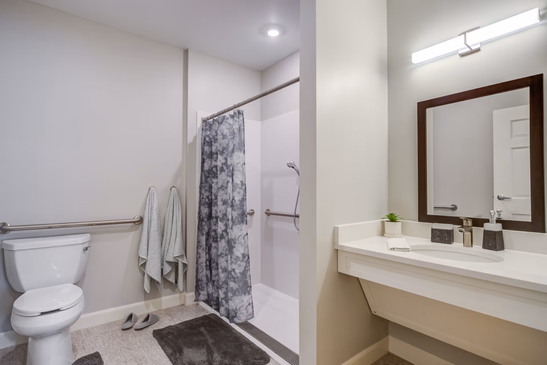 A clean and modern bathroom with a white toilet, two hanging towels, a walk-in shower with a gray floral shower curtain, a dark gray bath mat, a white vanity with a sink, a small potted plant, soap dispensers, a large mirror with a dark wooden frame, and a bright horizontal light fixture above the mirror.