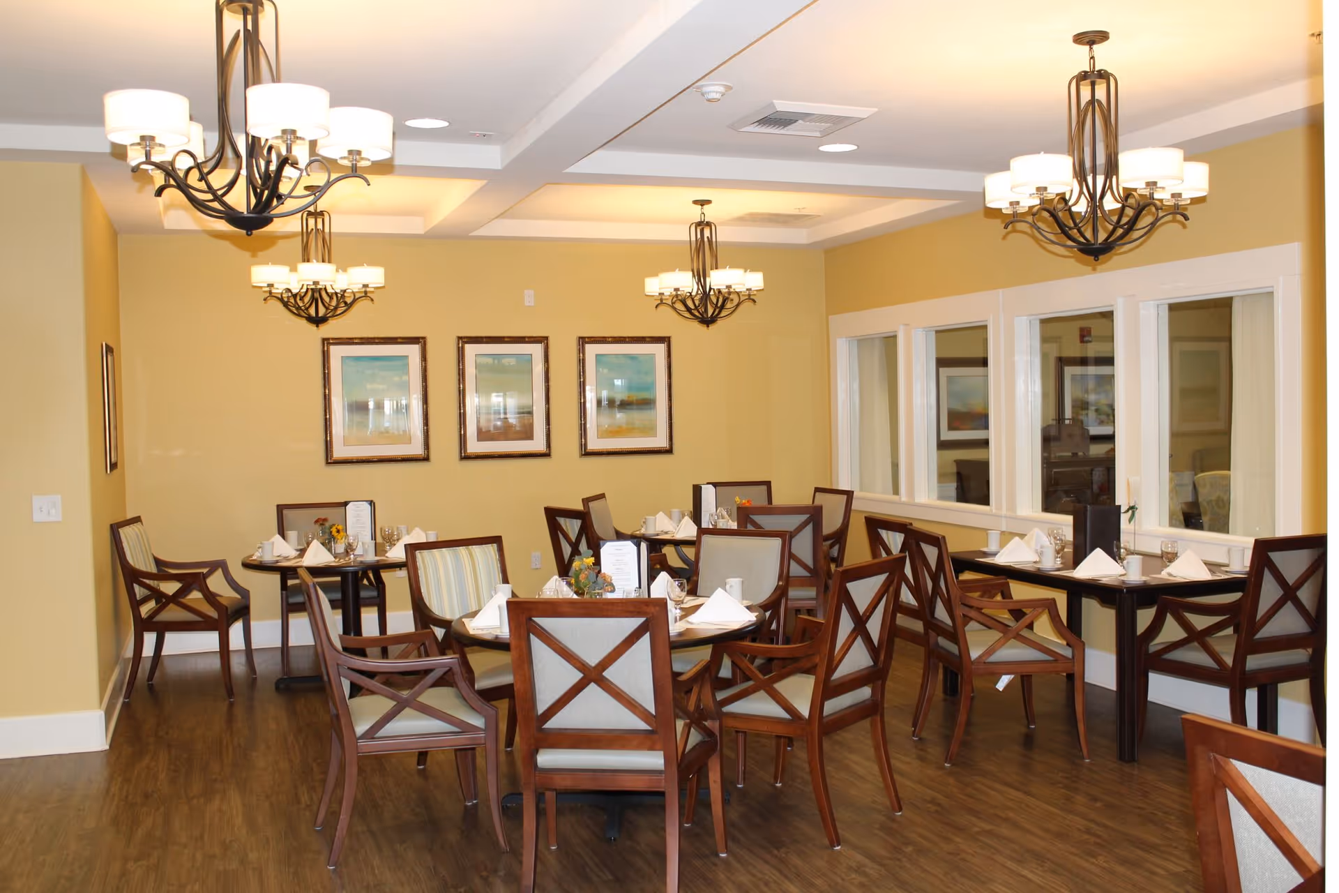 A dining room with several wooden tables and chairs arranged neatly. Each table is set with white napkins, glasses, and small flower arrangements. The walls are painted yellow and decorated with framed artwork. Multiple chandeliers with white lampshades hang from the ceiling, providing warm lighting. There are windows on one side of the room.