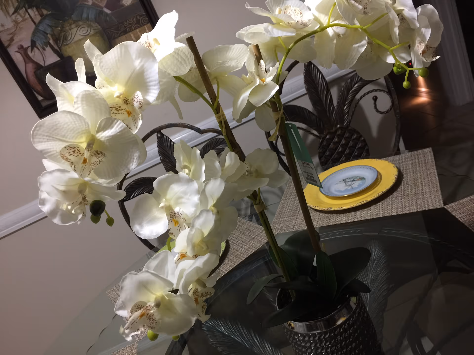 Close-up of a white orchid plant in a decorative pot placed on a glass dining table with woven placemats. Behind the table are metal chairs with pineapple-shaped backrests. A framed painting is visible on the wall in the background.