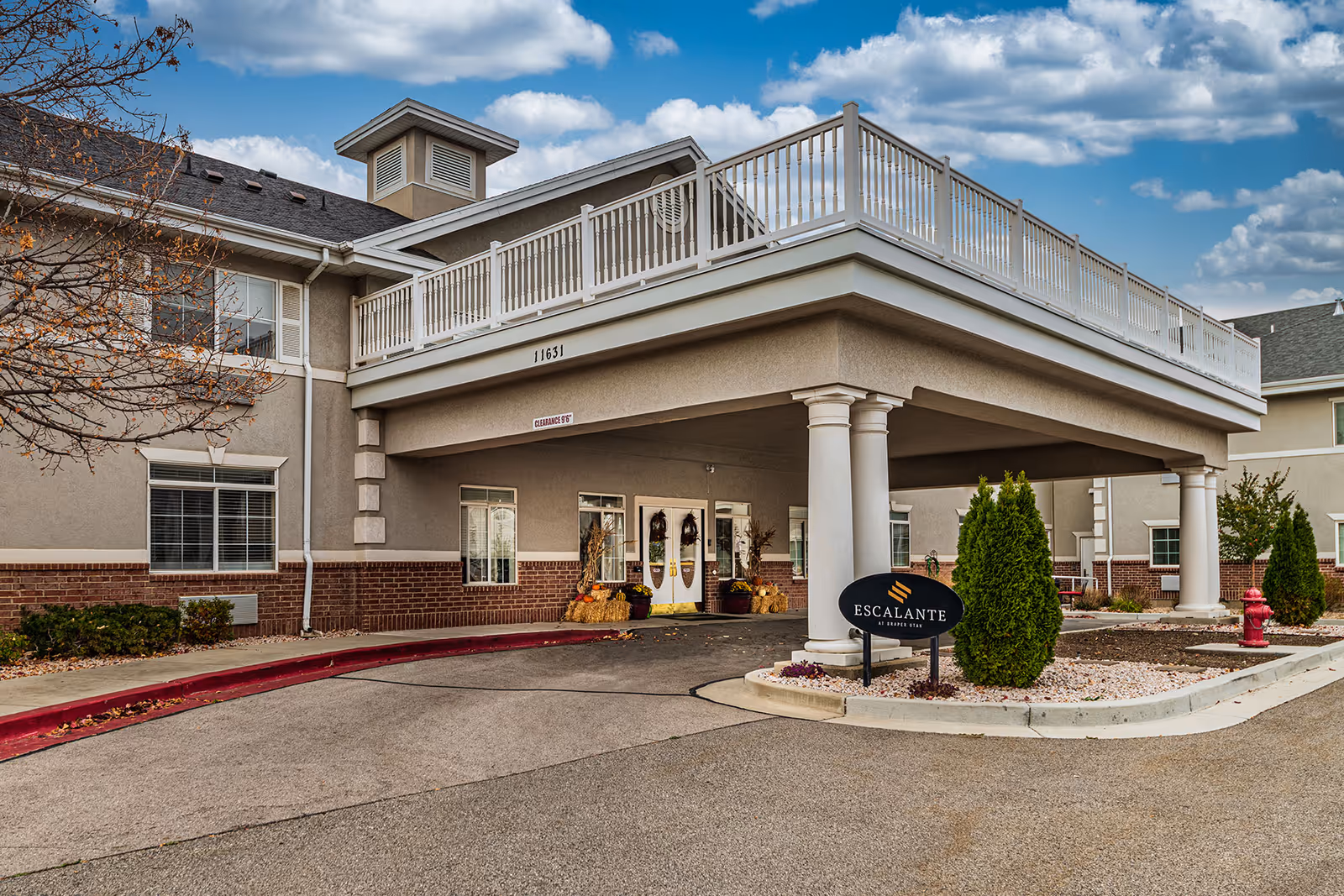 Exterior view of a senior living facility entrance with a covered drop-off area supported by columns. The building has beige and brick walls, white railings on the upper balcony, and a sign reading 'Escalante Senior Living' near a small landscaped area with bushes and a fire hydrant. The sky is partly cloudy.