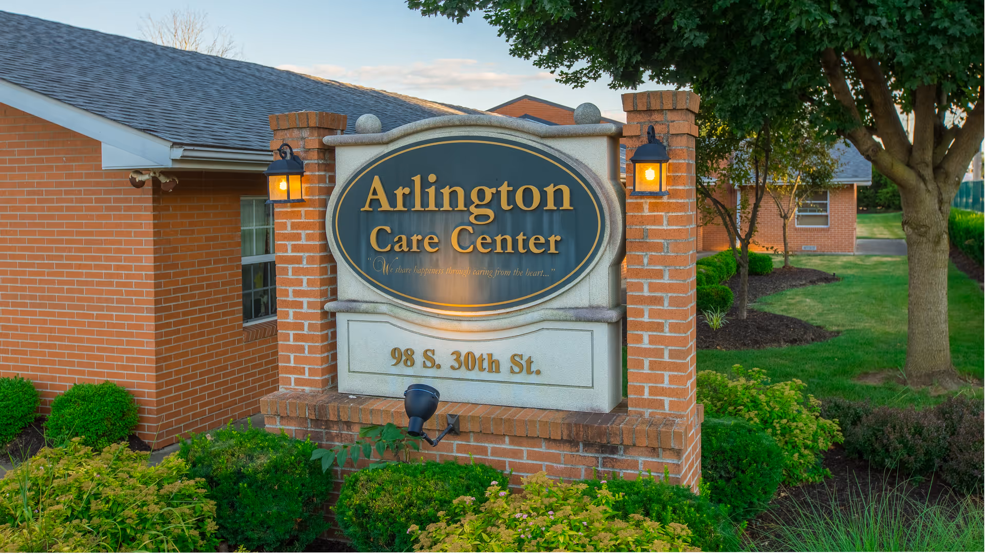 Outdoor view of a brick sign for Arlington Care Center located at 98 S. 30th St., surrounded by green bushes and trees with a building in the background.