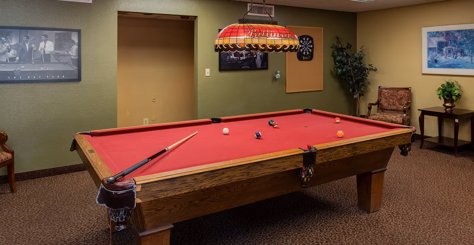 A red-felt pool table in a carpeted game room with a hanging billiards lamp, cue stick, chairs, dartboard, and framed wall art.