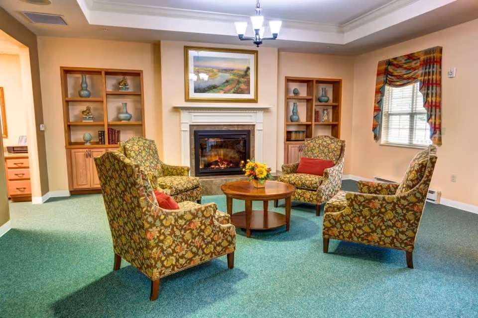 A cozy living room with four floral-patterned armchairs arranged around a round wooden coffee table with a flower arrangement. The room features a fireplace with a framed landscape painting above it, built-in wooden shelves with decorative items, a window with colorful curtains, and green carpeted flooring.