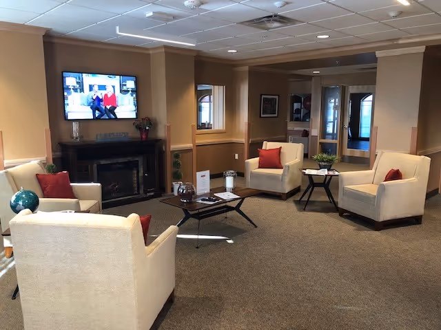 Common room with beige armchairs and red pillows arranged around coffee tables, a fireplace and a wall-mounted TV.