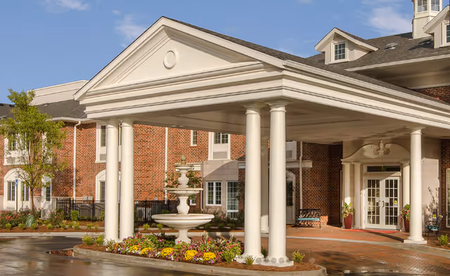 Brick senior living facility with a white portico, columns, fountain, and landscaped entrance.