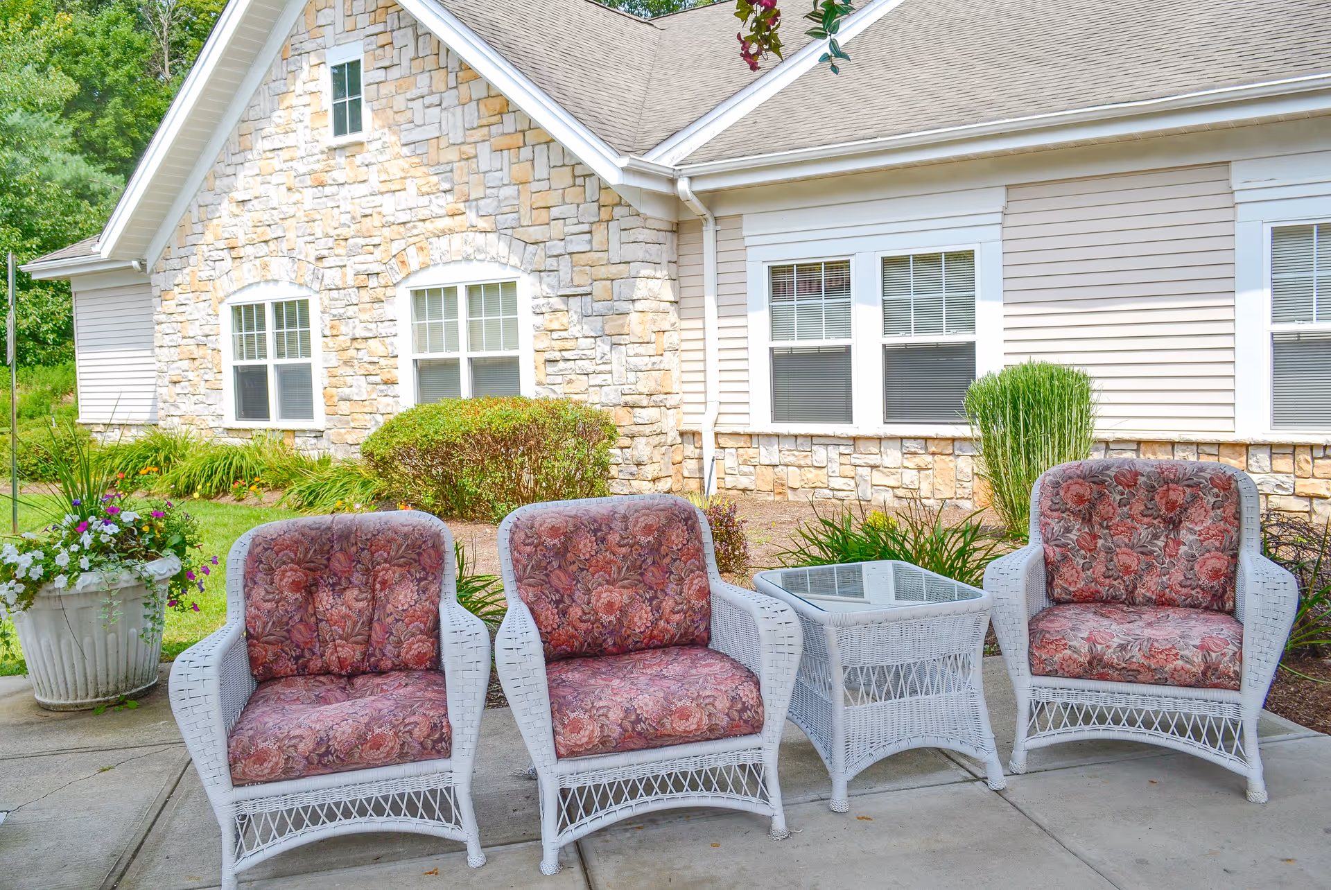 Outdoor patio area with three white wicker chairs featuring floral cushions and a matching wicker side table with a glass top, set on a concrete surface in front of a building with stone and siding exterior walls and multiple windows. There is greenery and a large planter with flowers nearby.