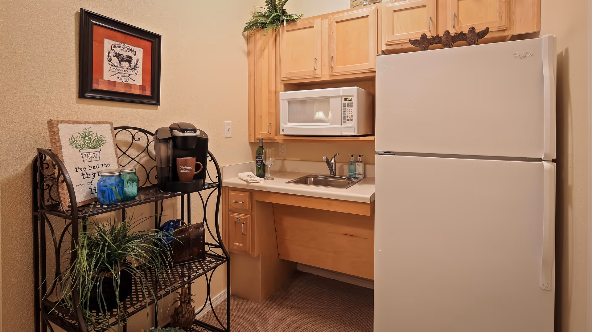 Small kitchen area with a white refrigerator, a microwave above the sink, wooden cabinets, and a black metal shelf holding a coffee maker, mugs, plants, and decorative items. A framed picture hangs on the wall above the shelf.