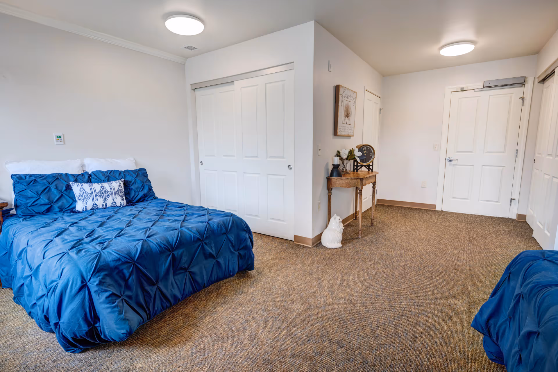 A bedroom with a bed covered in a blue comforter and matching pillows. The room has beige carpet flooring, white walls, and a white closet with sliding doors. There is a small wooden table against the wall with decorative items including a clock, a vase with flowers, and a small sculpture of a dog on the floor beside it. The room is well-lit with ceiling lights.