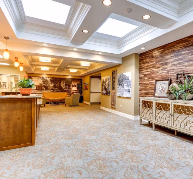 A spacious and well-lit interior common area in a senior living facility with skylights, recessed lighting, and pendant lights. The room features a patterned carpet, a wooden reception desk with a potted plant, a seating area with chairs and a sofa, and decorative wall art including a large black and white mural and framed pictures. There is also a wooden sideboard with plants and decorative items against a textured wooden accent wall.