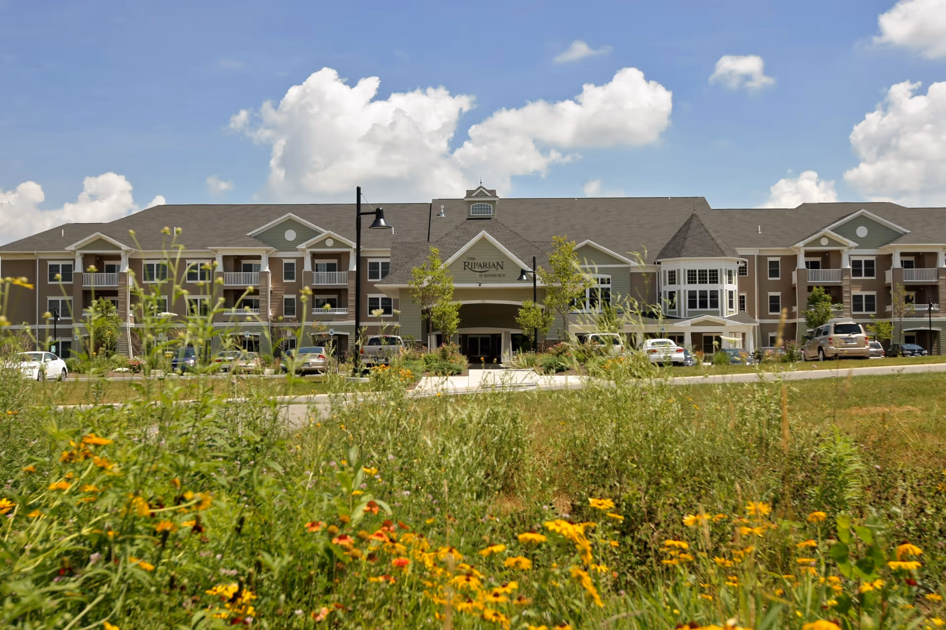 Front exterior view of a large senior living facility named The Riparian at Rivers Run, with a well-maintained garden and wildflowers in the foreground, several cars parked near the entrance, and a partly cloudy blue sky above.