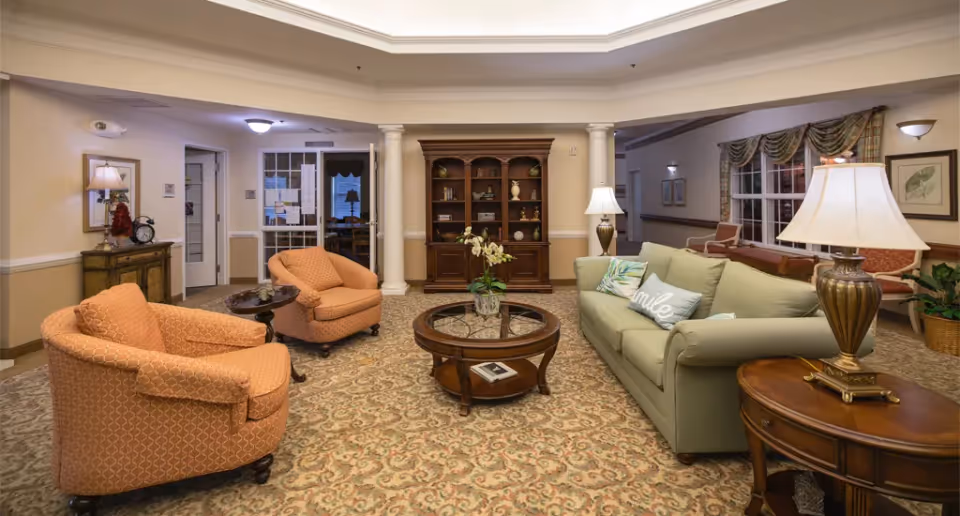 A cozy living room area in a senior living facility with a green sofa adorned with decorative pillows, two orange upholstered armchairs, a round wooden coffee table with a glass top, and a wooden side table with a lamp. The room features a patterned carpet, a wooden bookshelf with decorative items, and soft lighting from lamps and wall sconces. There are windows with draped curtains and a small table with chairs in the background.