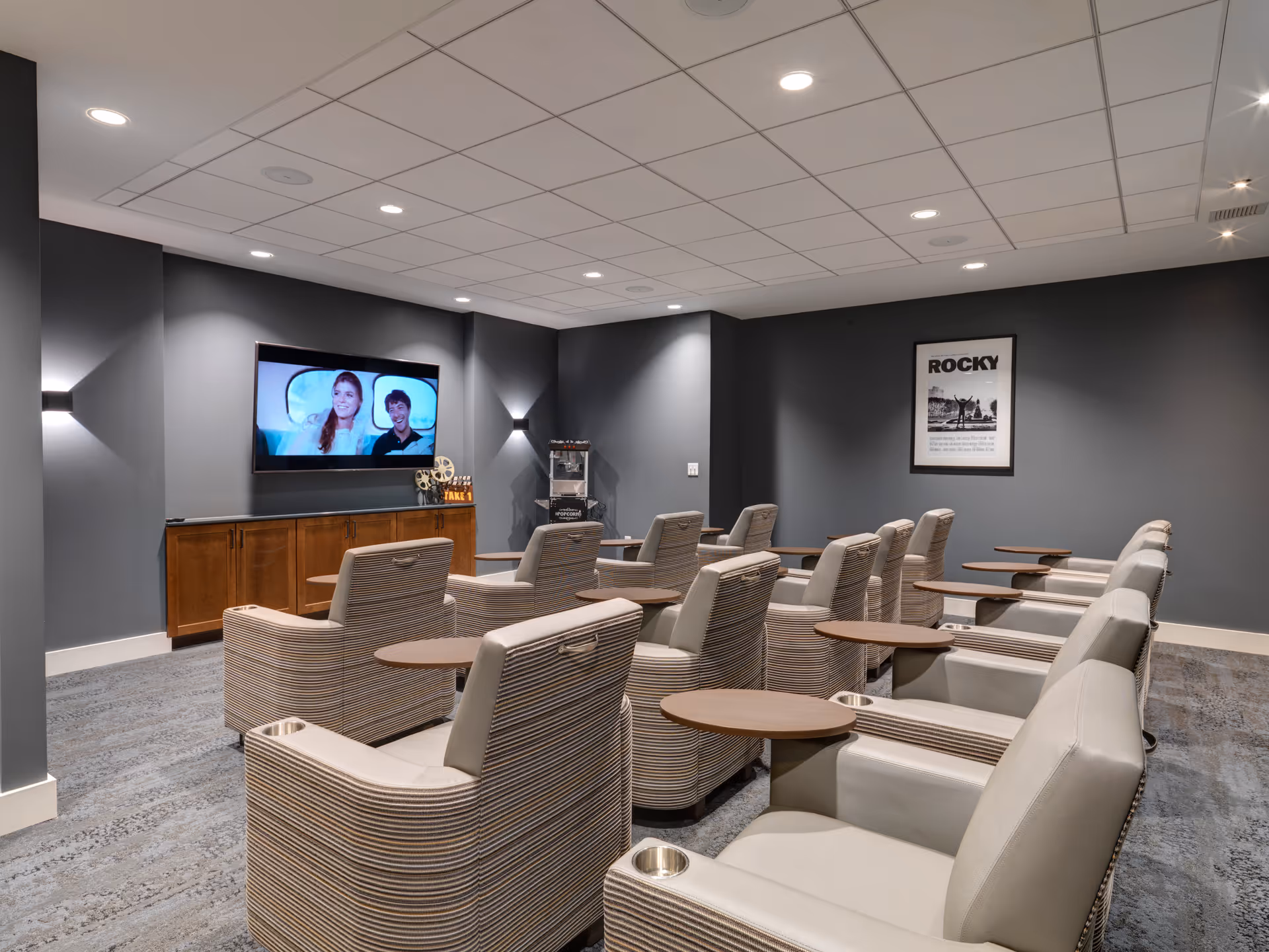 A modern movie theater room with multiple rows of comfortable beige and striped armchairs, each equipped with a small wooden table and cup holder. A large flat-screen TV is mounted on a gray wall, showing a scene with two people. Below the TV is a wooden cabinet, and a popcorn machine is placed in the corner. A framed poster of the movie Rocky hangs on the wall.