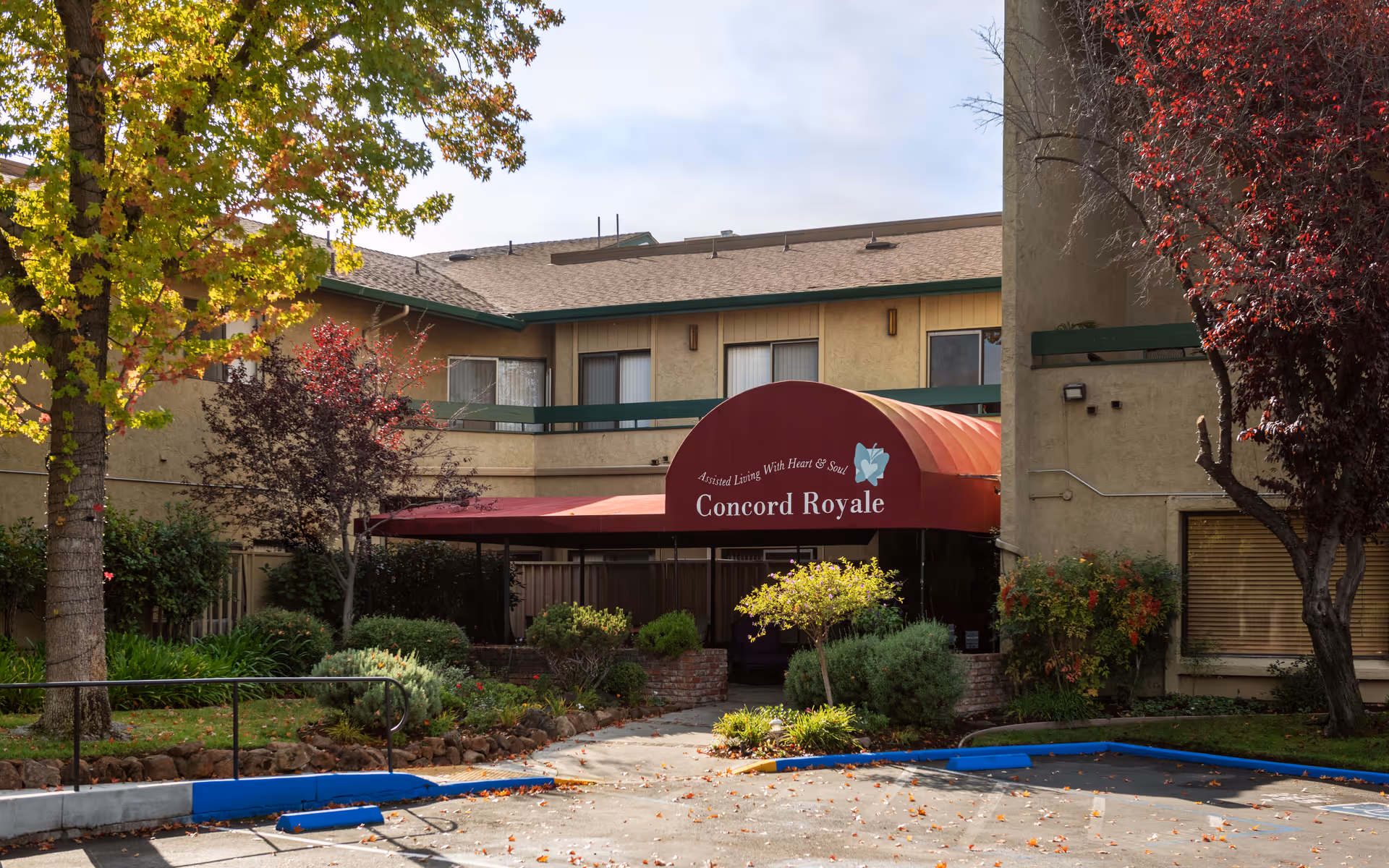 Exterior view of Concord Royale assisted living facility entrance with a red canopy that reads 'Assisted Living With Heart & Soul Concord Royale'. The building is surrounded by trees and shrubs with a parking area in front.