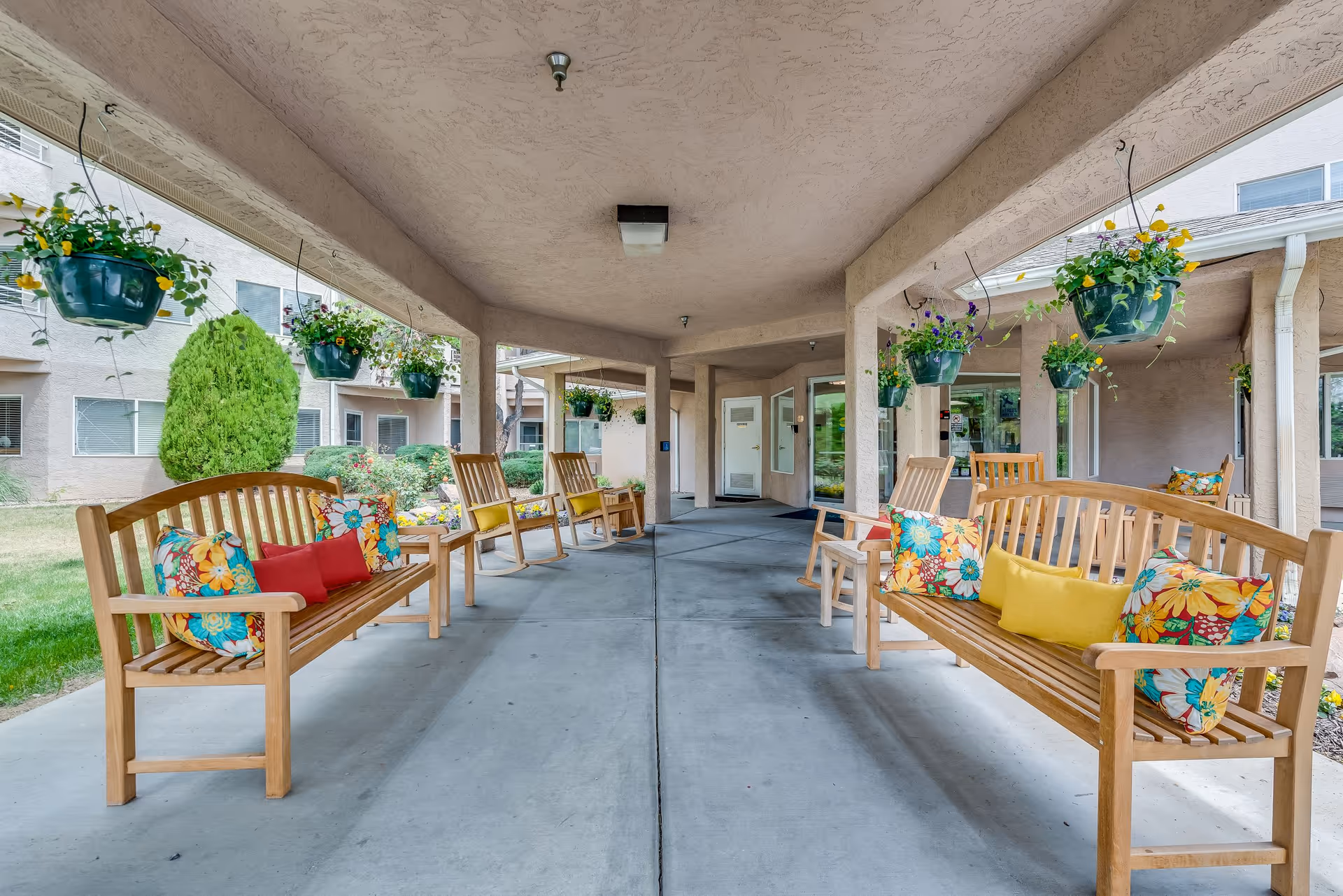 Covered outdoor seating area with wooden benches and rocking chairs adorned with colorful floral and solid pillows. Hanging flower pots with blooming plants are suspended from the ceiling. The area is part of a senior living facility with beige stucco walls and windows visible in the background.