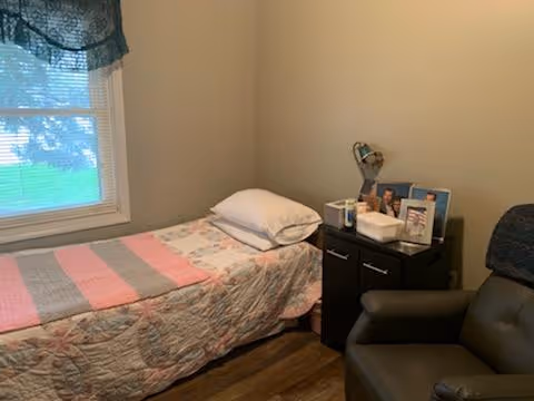A small bedroom with a single bed covered with a floral and striped quilt, a pillow, and a window with a blue valance. Next to the bed is a black nightstand with a lamp, tissue box, and framed photos. A dark-colored recliner chair is positioned nearby.