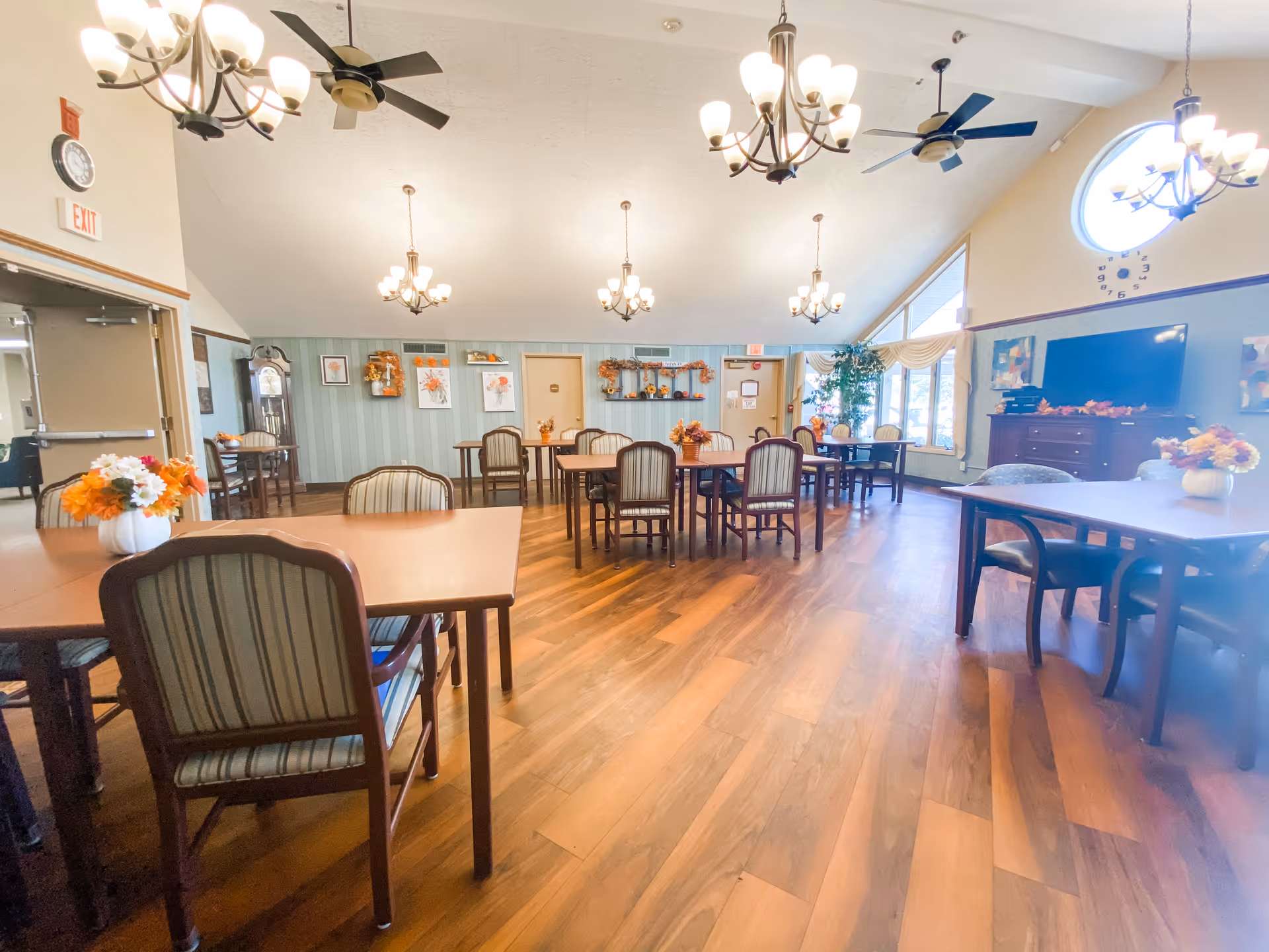 A spacious dining room with multiple wooden tables and chairs arranged neatly. The room has wooden flooring, several chandeliers hanging from the ceiling, ceiling fans, and large windows allowing natural light to enter. The walls are decorated with framed pictures and seasonal decorations. There is a large TV mounted on the wall and a grandfather clock near the entrance.