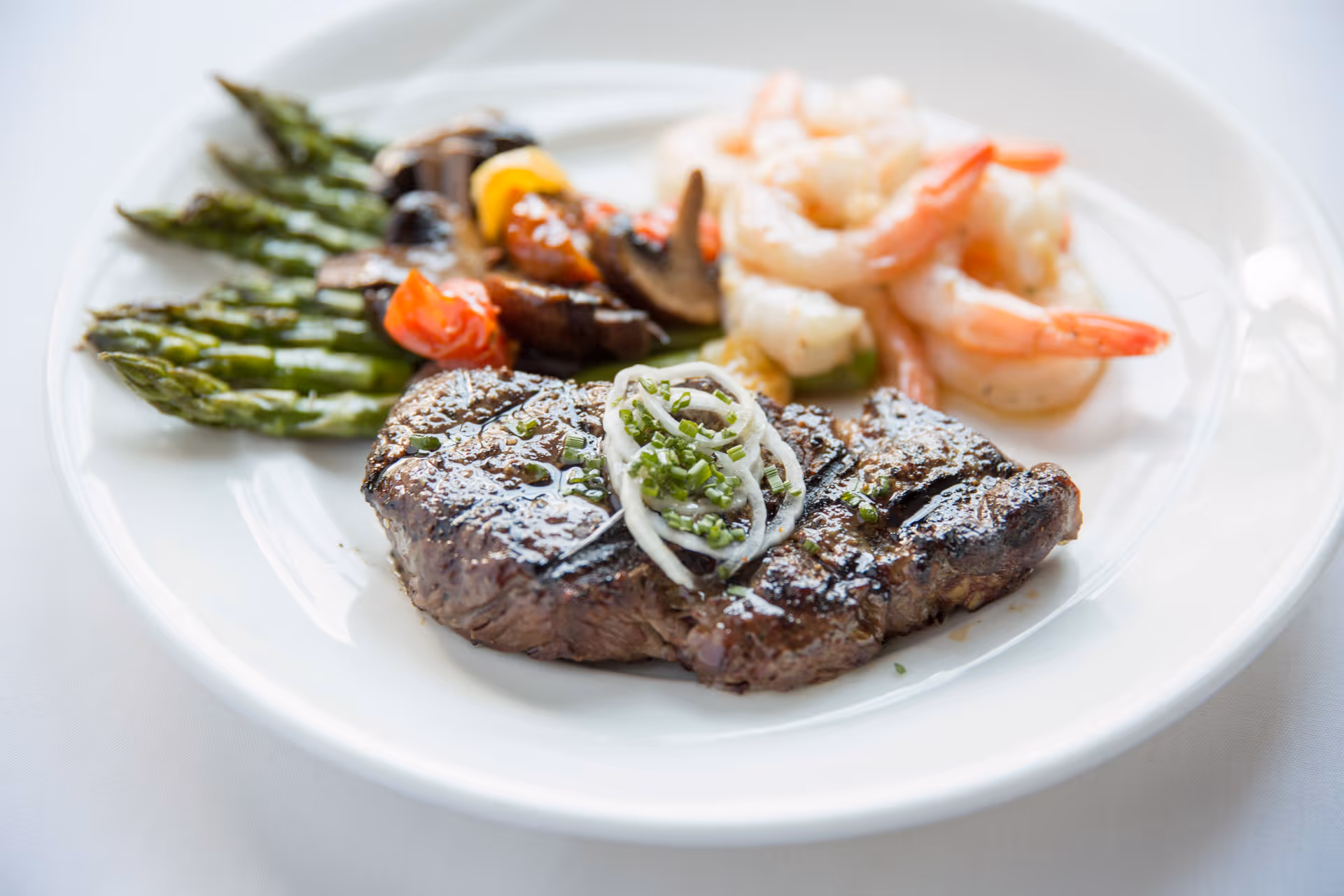 A white plate with a grilled steak topped with sliced onions and chives, served with grilled asparagus, sautéed mushrooms and cherry tomatoes, and cooked shrimp.