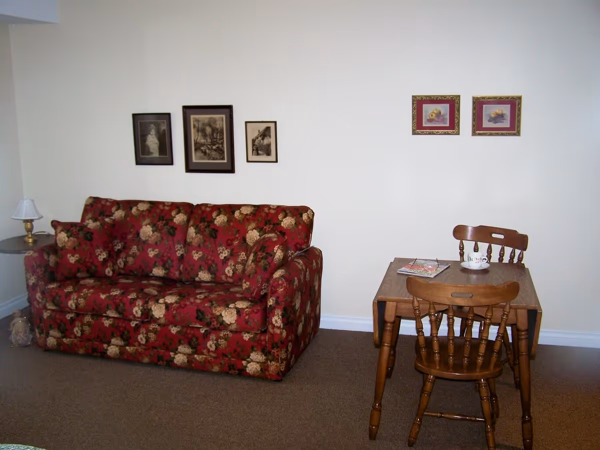 A small living area with a red floral patterned couch on the left and a wooden table with two matching chairs on the right. The table has a cup and a magazine on it. There are five framed pictures hanging on the white wall behind the furniture.