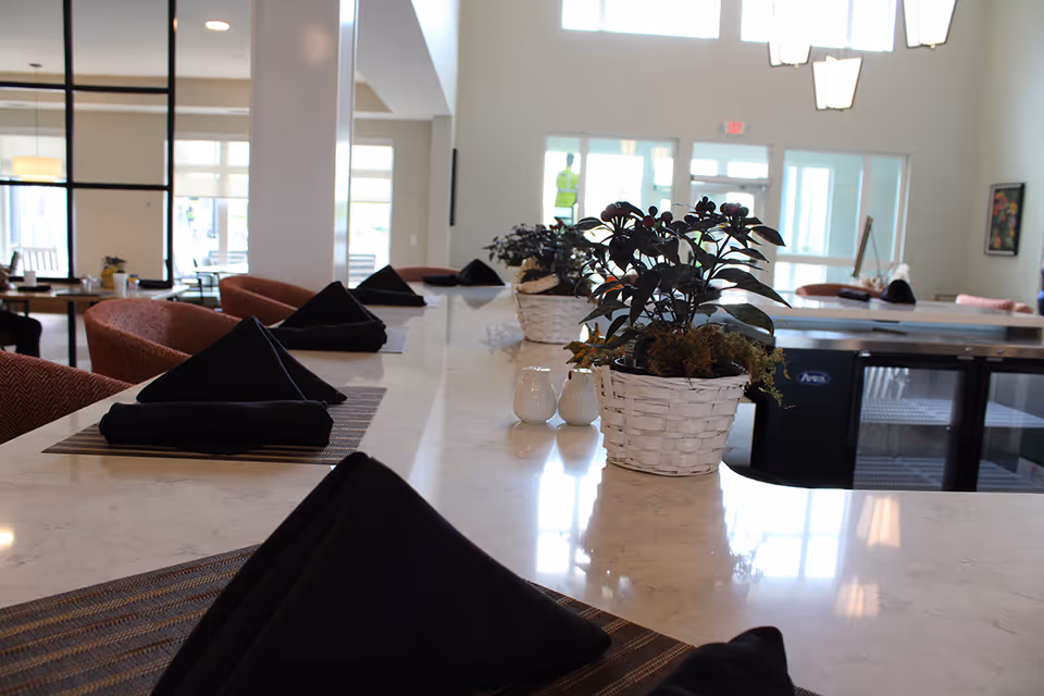 Bright communal dining counter with folded black napkins, potted plants, and seating in a senior living facility.