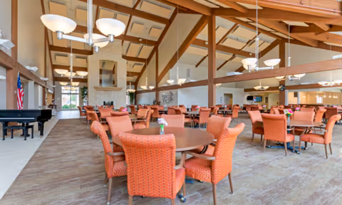 Spacious dining room with round tables and orange upholstered chairs under a high beamed ceiling and chandeliers.