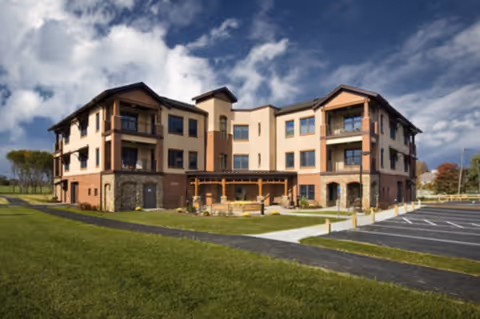 Exterior view of a three-story senior living facility building with a mix of brick and beige siding under a partly cloudy sky. The building has balconies, large windows, and a covered outdoor seating area. There is a paved parking lot and green lawn surrounding the building.