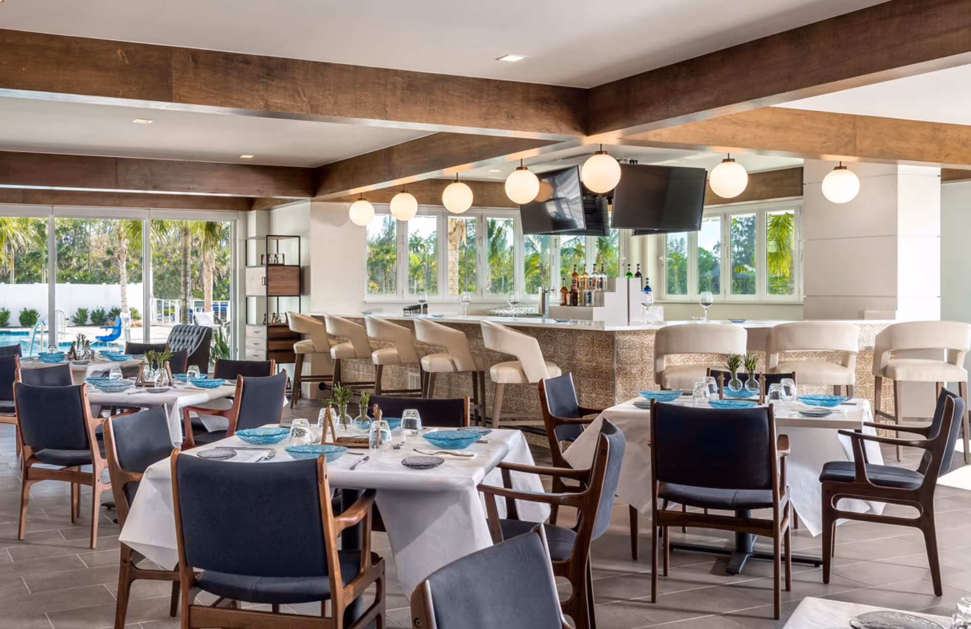 A bright and modern dining area with tables covered in white tablecloths, set with blue plates, glasses, and silverware. The room features dark blue cushioned chairs with wooden frames, a bar with beige bar stools, hanging globe lights, and large windows showing palm trees and an outdoor pool area.