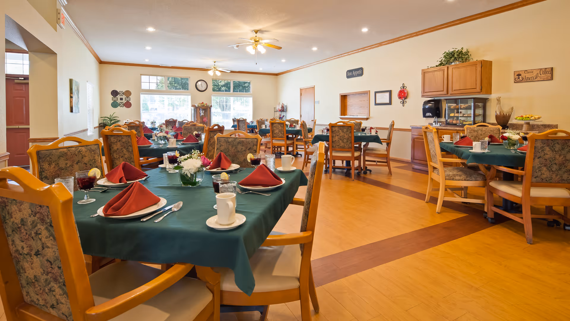 A bright dining room with multiple wooden tables covered with green tablecloths and set with white plates, cups, silverware, and red folded napkins. The room has wooden chairs with floral upholstery, large windows letting in natural light, and light-colored walls decorated with signs and plants. The floor is a combination of light and dark wood patterns.