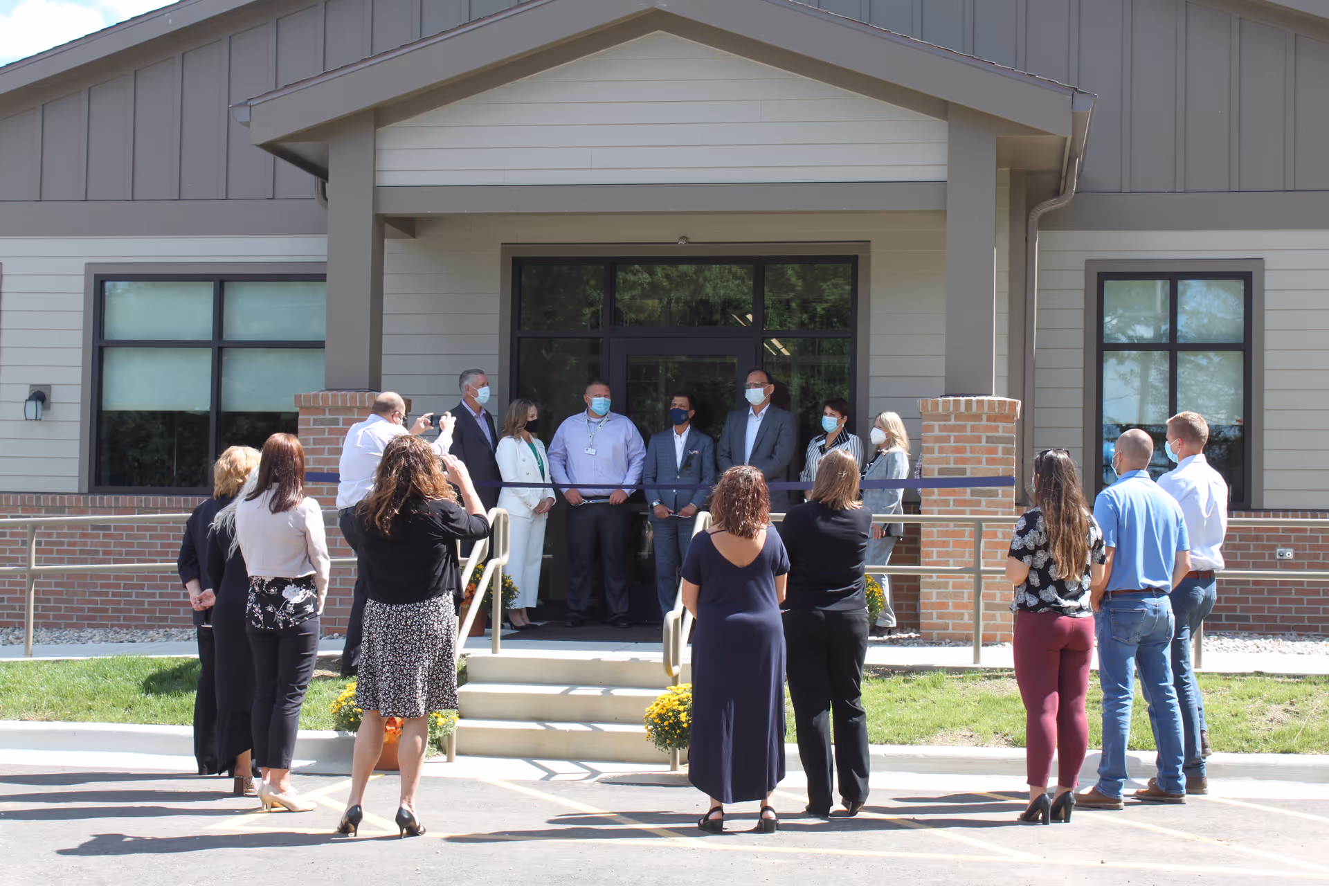 A group of people gathered outside the entrance of a building for a ribbon-cutting ceremony. Several individuals are standing on the steps holding a ribbon, while others stand facing them, some taking photos. Everyone is wearing face masks.