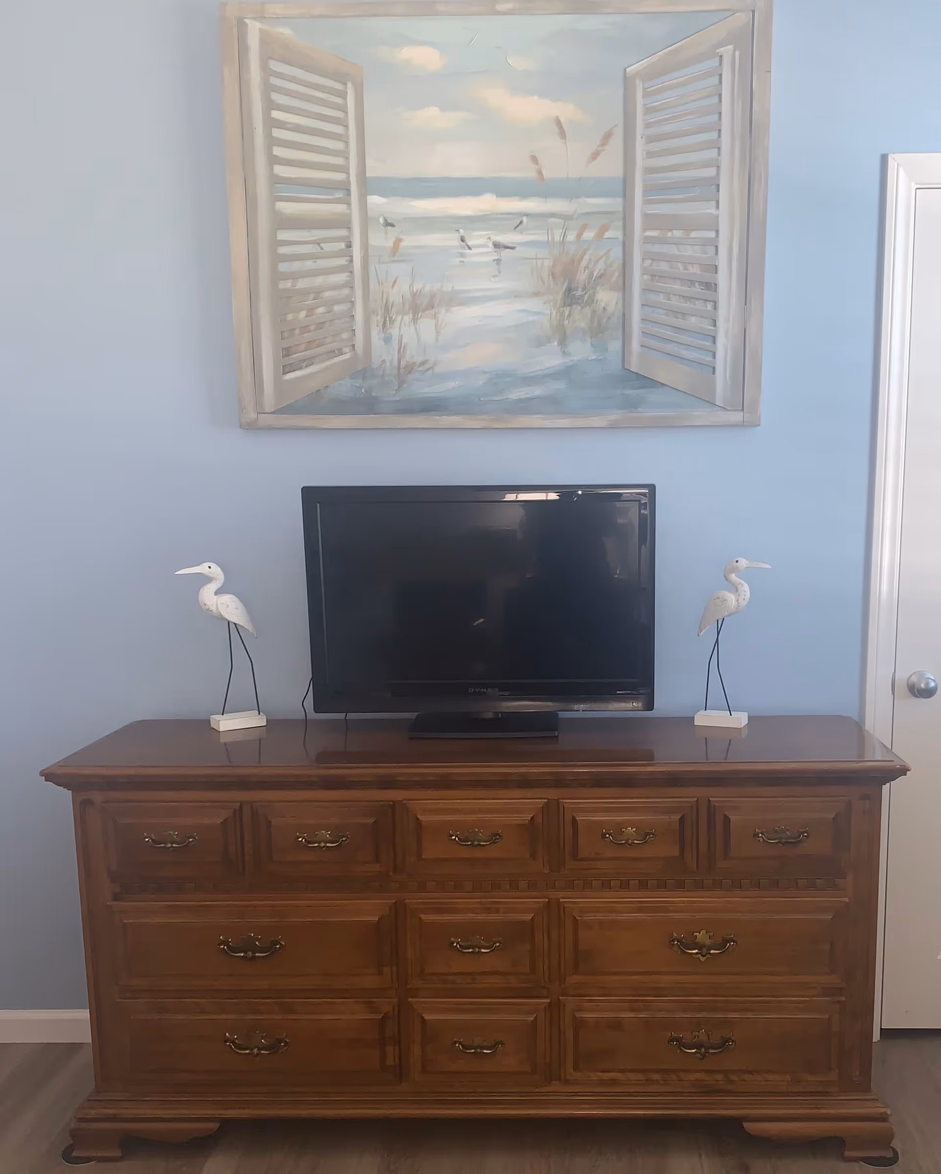 A wooden dresser with multiple drawers and brass handles, topped with a flat-screen TV and two white bird figurines on either side. Above the dresser is a painting depicting an open window with shutters looking out onto a beach scene with seagulls and grasses.