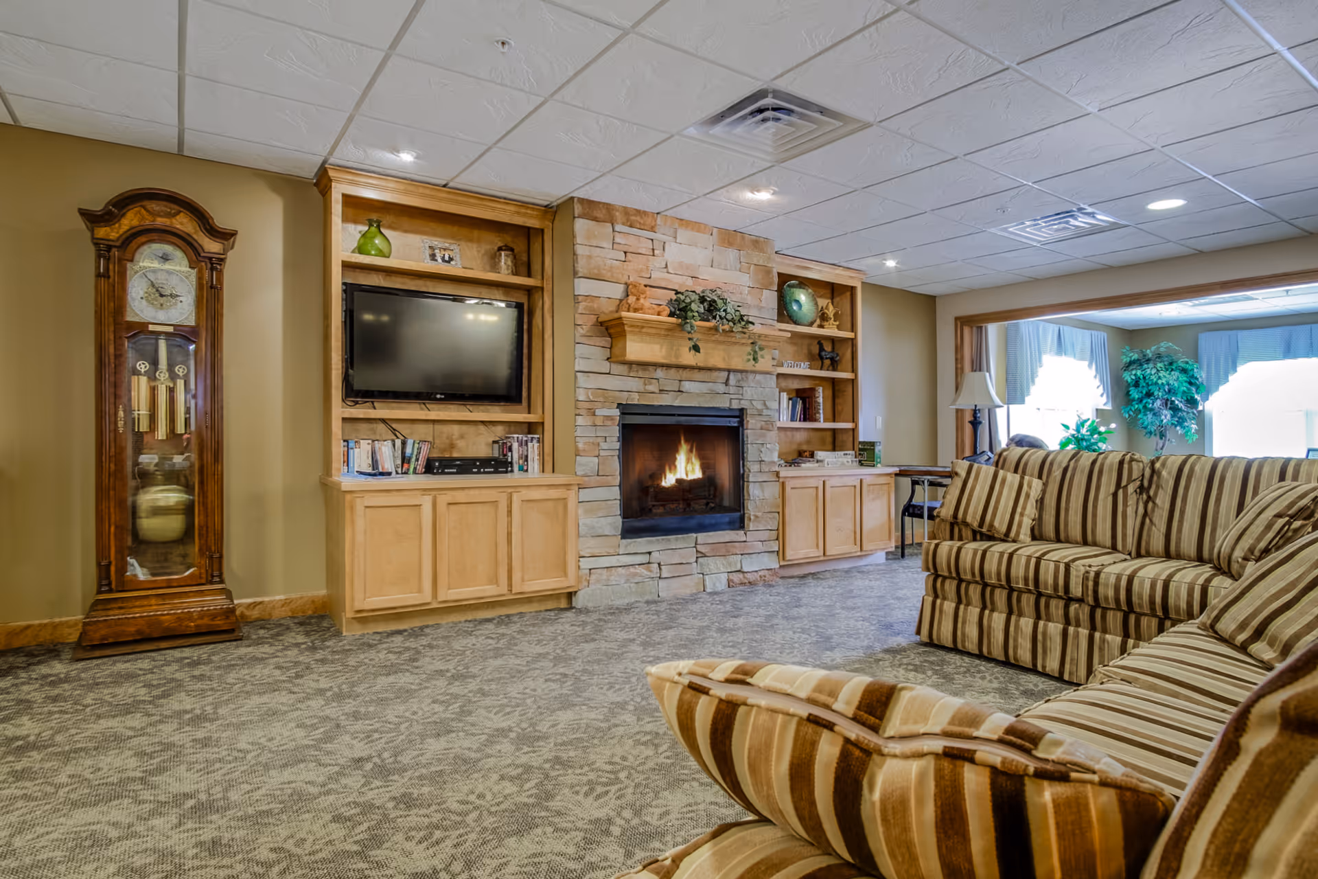 A cozy living room with a stone fireplace in the center, flanked by wooden built-in shelves and cabinets. A flat-screen TV is mounted on the left shelving unit. To the left of the fireplace is a tall wooden grandfather clock. The room has patterned carpet flooring and a striped sofa with matching cushions in the foreground. Large windows with curtains and potted plants are visible in the background.