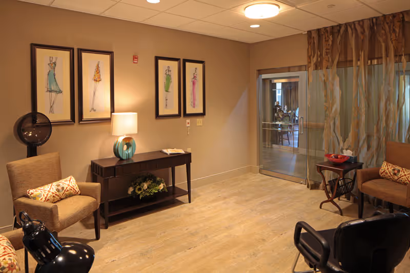 A cozy interior lounge area with two upholstered armchairs each with a decorative pillow, a dark wooden console table with a lamp and floral arrangement, four framed fashion sketches on the wall, a floor fan, and a glass sliding door with sheer curtains leading to another room with tables and chairs.