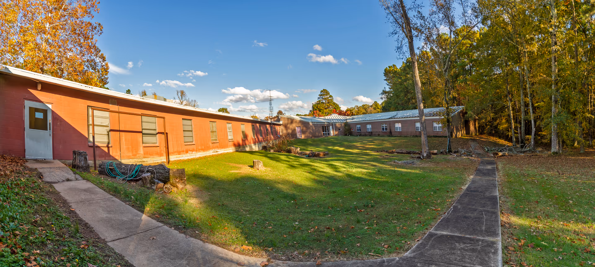 Exterior view of a single-story assisted living facility building with red brick walls and multiple windows, surrounded by a grassy lawn and trees with autumn foliage under a blue sky with scattered clouds. A concrete walkway leads to the entrance on the left side and another path extends to the right through the lawn.