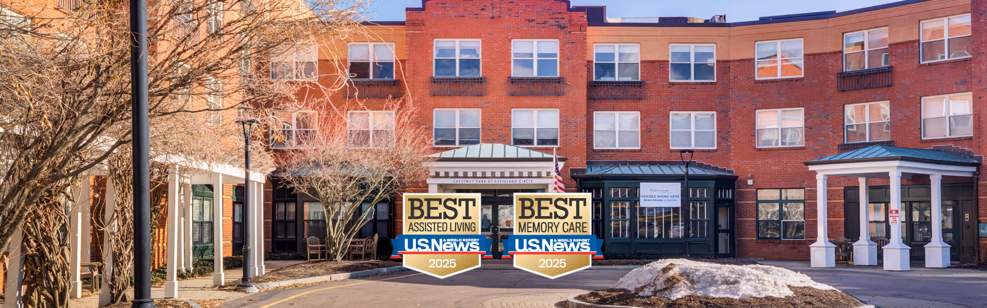 Red-brick senior living building front with a covered entrance and US News 'Best Assisted Living' and 'Best Memory Care' 2025 badges displayed.