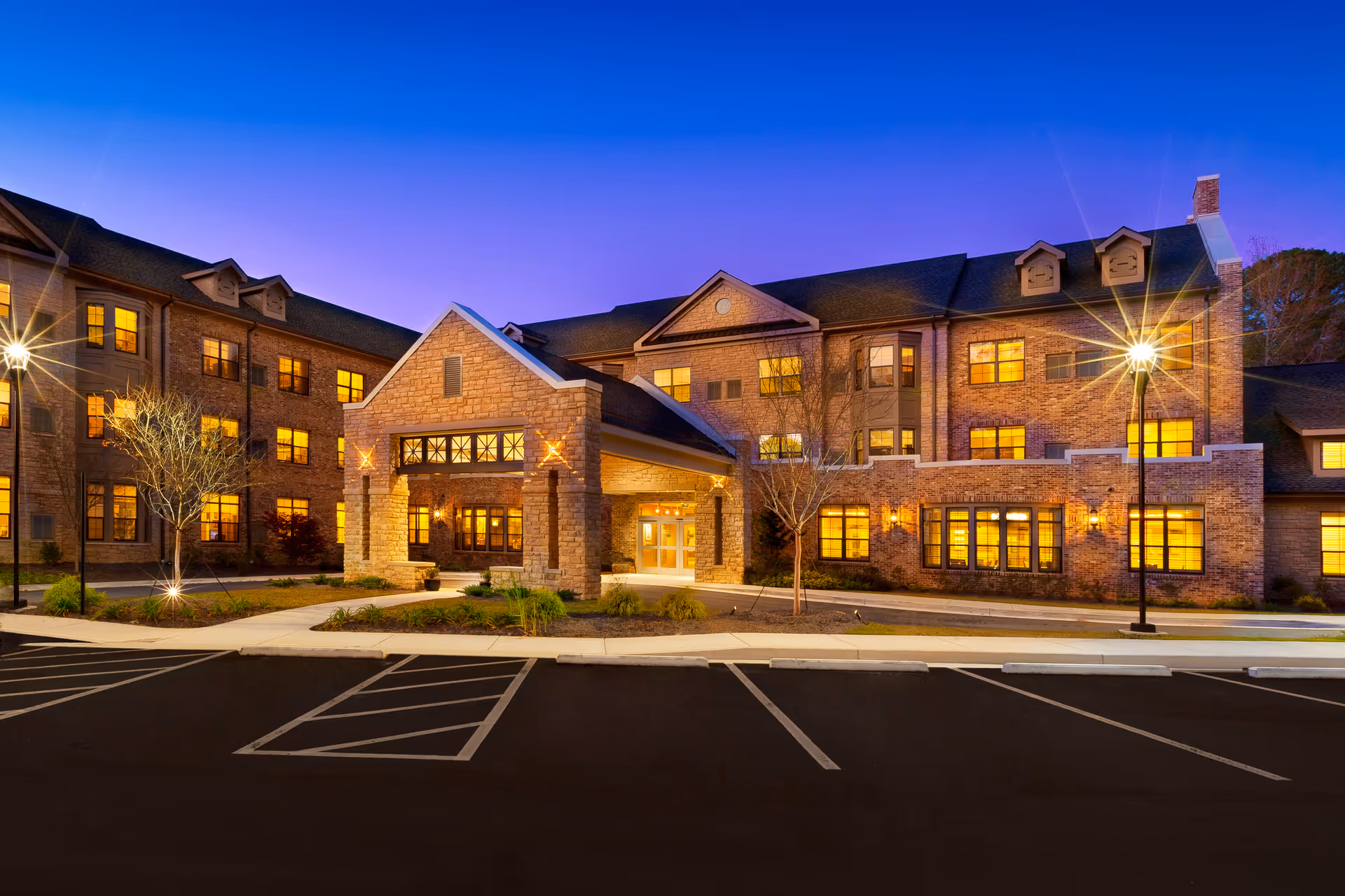 Exterior view of Berman Commons senior living facility at dusk, showing a large brick building with multiple windows illuminated from inside, a covered entrance with stone pillars, landscaped greenery, and a parking lot in the foreground.