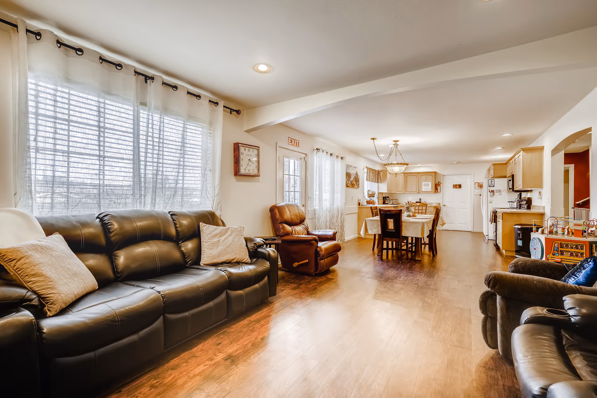 A bright and spacious living area with a black leather sofa and recliner chairs, a dining table with chairs, and a kitchen with wooden cabinets in the background. Large windows with sheer white curtains allow natural light to fill the room. The floor is wooden, and there is a clock and an exit sign on the wall.