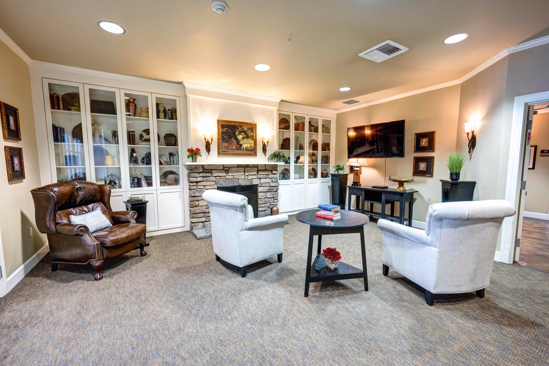 A cozy living room area with a stone fireplace flanked by white built-in cabinets filled with decorative items and books. The room features a brown leather armchair, two light gray upholstered armchairs around a round black coffee table with books and a flower vase. A flat-screen TV is mounted on the wall above a black console table with a lamp and decorative items. The walls are beige with framed artwork and wall sconces providing warm lighting.